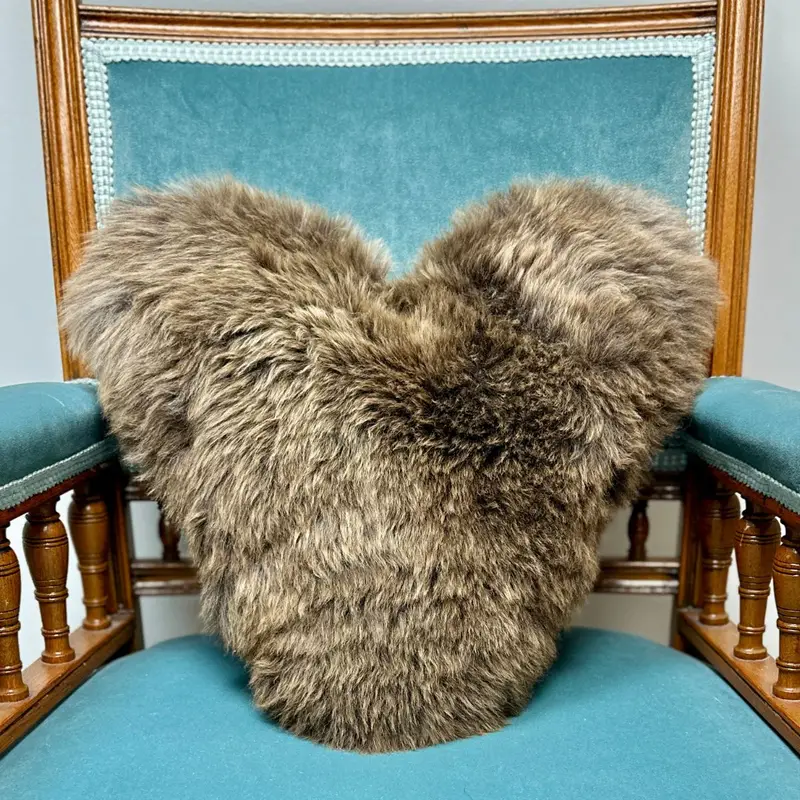 Natural British rare breed heart shaped sheepskin cushion placed on a deep blue velvet chair in a cosy interior setting.