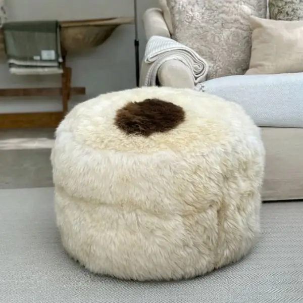 A very large ivory rare breed sheepskin pouffe with a cute chocolate patch in a neutral coloured room in front of a large sofa.