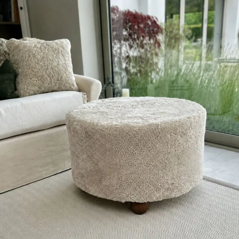 Extra large oatmeal shearling sheepskin stool with walnut hardwood barrel feet used as a coffee table in front of a beige sofa