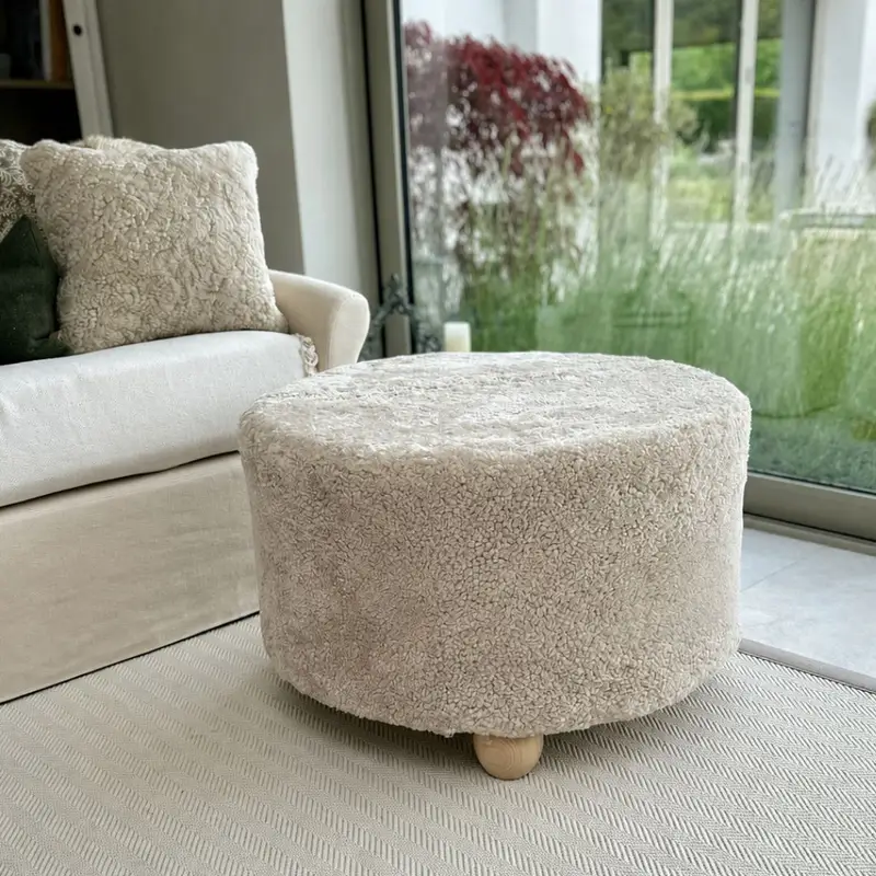 Extra large oatmeal shearling sheepskin stool with natural hardwood barrel feet used as a coffee table in front of a beige sofa